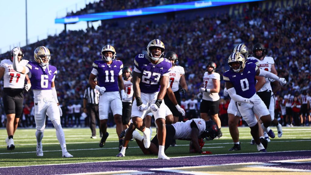 A football player in a purple jersey with the number 23 celebrates a touchdown while teammates and opponents are visible on the field during a game.
