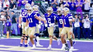 A group of football players in purple and gold uniforms walk across the field, with spectators and photographers visible in the background.